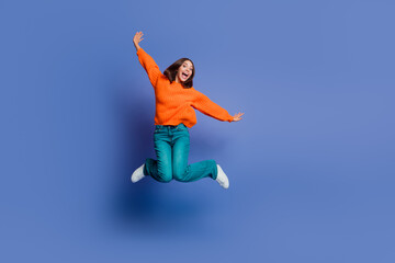 Young woman in casual orange sweater, jumping with excitement against a blue background showcasing...