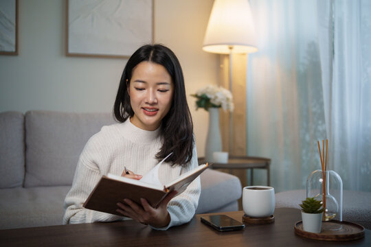 Young Asian woman sitting at a table, smiling while writing in a notebook at home.