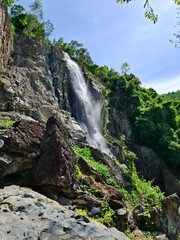 High waterfall flowing down the rocky cliff surrounded by green tropical forest and sunlight in Vietnam. Beautiful natural landscape with fresh air and bright summer sky.