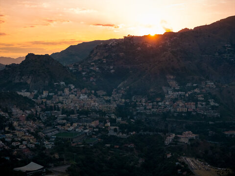 Aerial view of the sun's radiant burst over mountain peaks and clustered buildings, Taormina, Sicily, Italy.