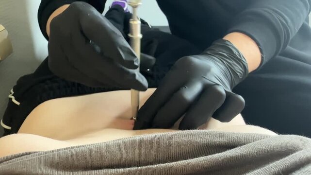 A female with light skin receives a navel piercing. A gloved hand holds a piercing tool near her abdomen. The setting is a tattoo studio.