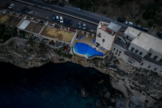 Aerial view of a striking blue swimming pool clinging to the rugged coastline next to buildings with parked cars, Taormina, Sicily, Italy.