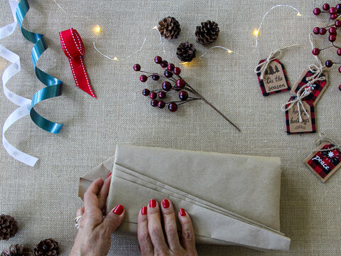 Flat lay overhead of woman wrapping Christmas gifts with Christmas supplies to pack Holiday presents on textured neutral surface using recycled kraft Paper.