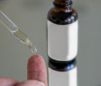 Closeup view of woman using a dropper to place a drop of serum on her fingertip and a glass serum bottle with white mockup friendly label in the background for skincare concept. 