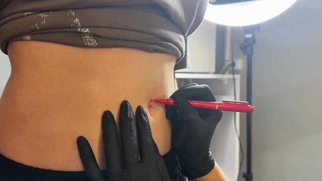 A young Caucasian woman with long dark hair is getting a navel piercing. A professional piercer in black gloves marks the spot with a red pen.