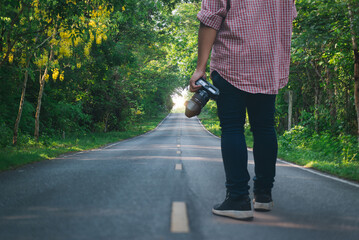 A person holding a camera stands on a quiet forest road, surrounded by lush green trees and morning sunlight ahead.
