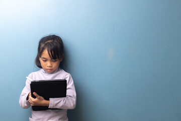 Asian little girl using digital tablet with stylus for online learning on blue wall background, concept of digital education, smart kid, and e-learning technology.