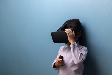 Asian little girl wearing VR headset and holding controller to explore virtual reality on blue wall background, concept of metaverse, digital education, innovation, and future technology.