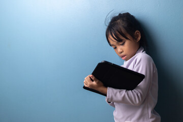 Asian child concentrating on digital tablet for online learning on blue wall background, concept of education technology, smart student, digital classroom, and e-learning lifestyle.