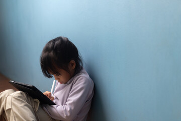 Asian child sitting on floor and focusing on tablet with stylus for online learning on blue wall background, concept of digital education, smart learning, and homeschooling technology.