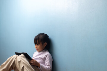 Asian little girl sitting and learning with digital tablet on blue wall background at home, concept of calm education, digital lifestyle, homeschooling, and smart learning technology.