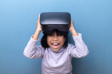 Amazed Asian kid with an excited face holding a VR headset. Happy child experiencing virtual reality, metaverse technology, and gaming on a blue background with copy space.
