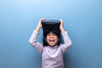 Happy Asian girl laughing with excitement while holding a VR headset. Child enjoying a fun virtual reality experience, metaverse gaming, or ed-tech on a blue background.