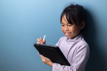 Smiling Asian child using digital tablet with stylus for online learning on blue wall background, concept of happy education, creative learning, and digital technology for kids.
