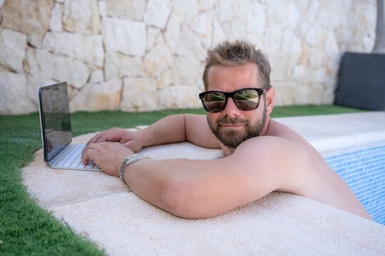 Portrait of a young bearded man wearing sunglasses in swimming pool while using tablet in summer time. Holidays concept.