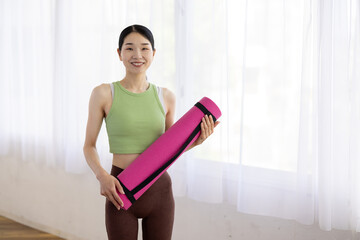 Smiling Asian Middle-Aged Woman in Activewear Holding Yoga Mat Indoors