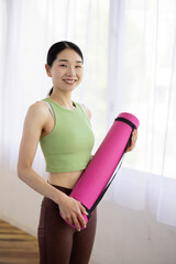 Smiling Asian Middle-Aged Woman in Activewear Holding Yoga Mat Indoors