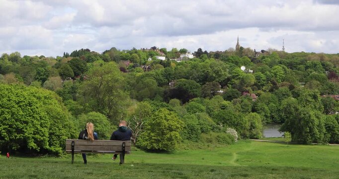 Couple sitting on a bench facing a lush valley of trees and rooftops on a bright spring day. Quiet pause, shared moment, nature and city meeting at the edge.