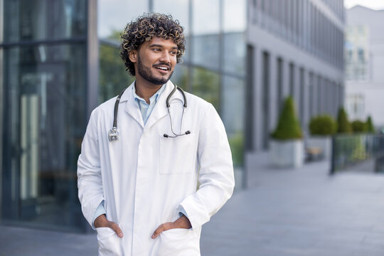 Smiling young Indian man stent, doctor standing in nephrectomy on the street near the clinic, holding hands in pockets and looking to the side