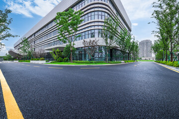 Empty asphalt road and entrance to a modern office building exterior with a glass facade and green trees.