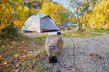 A power bank charges a phone outdoors against the backdrop of a tent and an autumn landscape.