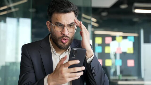 Upset worried businessman in formal suit reads bad news on phone sitting at workplace in business office. Frustrated entrepreneur is sad while reviewing negative information on a smartphone. Close up