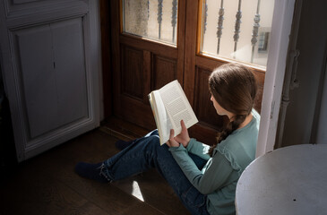 Preteen girl reading a book by a sunlit window, sitting on wooden floor. Calm indoor moment showing childhood, education and slow lifestyle at home.