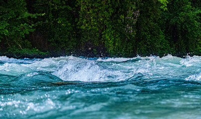 Close-up of fast flowing river with splashing water and green vegetation background. Showing natural motion, freshness and energy of wild stream in nature. Selective focus.