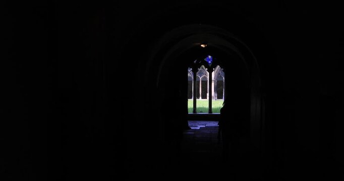 People in silhouette walk under a dark stone archway, facing a bright cloister garden and stained glass. Contrast of shadow and light suggests contemplation, solitude, and inner reflection.