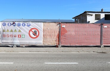 Temporary construction fence with  information board depicting the warning, prohibition and mandatory signs for the building activity. Street in front, building on behind. Background for copy space.