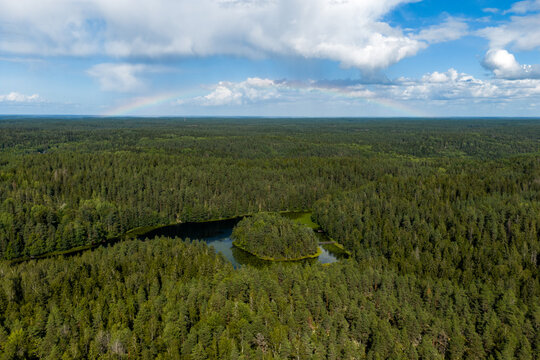 Aerial view of a serene lake nestled within a dense, verdant forest under a sky brushed with soft clouds and a faint rainbow, Espoo, Uusimaa, Finland.