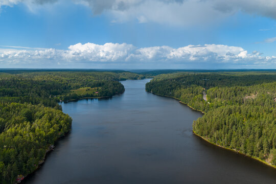 Aerial view of a vast, dark river winds through a dense forest, its waters reflecting the sky, contrasting with the vibrant green canopy, Espoo, Uusimaa, Finland.