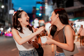 Two friends happily sharing street food together at a night market, showing asian cuisine, travel lifestyle, joyful social interaction, friendly atmosphere and fun candid moments outdoors in the city