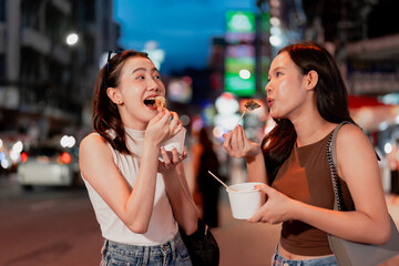 Two friends eating delicious street food together at a night market, showing asian cuisine, travel lifestyle, friendly atmosphere, candid happy emotions and fun social moments outdoors