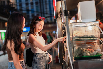 Two friends ordering street food at a night market stall, showing Asian cuisine, tourism lifestyle, cultural experience, casual social atmosphere and fun candid moments while exploring outdoors