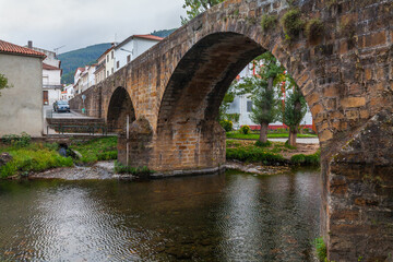 An ancient, moss-covered stone arch bridge spans a calm river in a quaint village