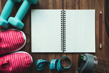 Top view of open notebook with pencil, pink sneakers, blue dumbbells, water bottle and measuring tape on wooden background with copy space, fitness planning and workout equipment flat lay