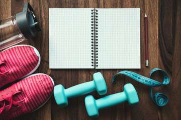 Top view of open notebook with pencil, pink sneakers, blue dumbbells, water bottle and measuring tape on wooden background with copy space, fitness planning and workout equipment flat lay