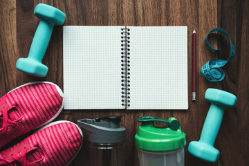 Top view of open notebook with pencil, pink sneakers, blue dumbbells, shaker, water bottle and measuring tape on wooden background with copy space, fitness planning and workout equipment flat lay