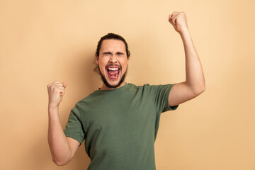 Energetic man celebrates victory with raised fists smiling wide green shirt beige background