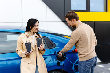 Young couple man and woman traveling by electric car having stop at charging station. Boyfriend plugging in cable to charge. Man talking with girlfriend, holding cup drinking hot coffee smiling