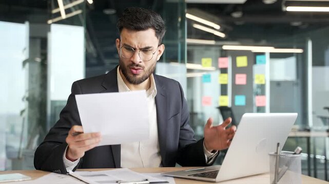 Confused businessman having difficulty with paperwork checking documents on laptop sitting at desk at workplace in business office. Puzzled entrepreneur does not understand the solution to the problem