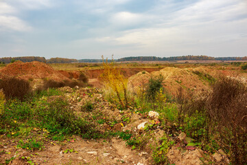 Autumn landscape of an open-pit quarry with reddish earth mounds and sparse vegetation under a cloudy sky. Distant forest line at the horizon