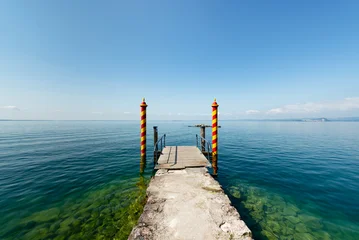 Fototapete Rund Pier Gelb-rot gestreifte Holzstangen am Bootsanleger der Locanda San Vigilio auf einer Halbinsel im Gardasee in der Morgensonne, Venetien, Italien  © Matthias Riedinger