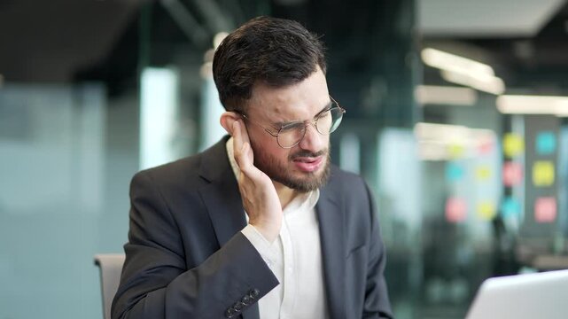 Businessman in formal suit suffers from ear pain sitting at workplace in office. Upset male touches his ear with his fingers. He has furuncle, shingles, eczema, ear diseases or otitis media. Earache