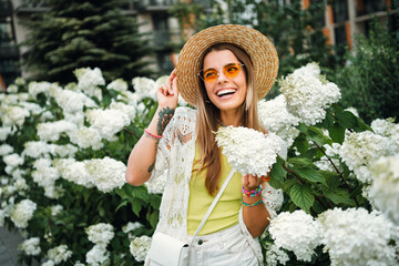 Happy young woman in sunhat among white hydrangeas smiles as she poses for a travel casual outdoor park shoot