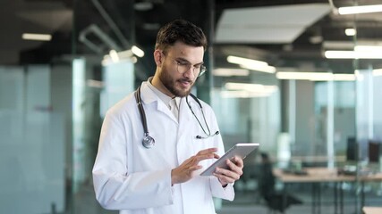 Portrait of serious handsome doctor in white coat working on digital tablet standing in modern hospital clinic looking at camera. Confident young focused medical worker physician in glasses in office - Powered by Adobe