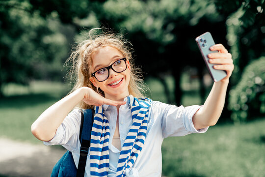 Young woman with glasses smiling and taking a selfie outdoors in casual attire enjoying a sunny day in a vibrant city