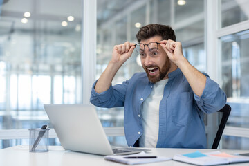 Man in an office setting expressing surprise and excitement, holding up his glasses, experiencing...
