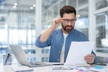 Obraz premium Young businessman adjusting spectacles while reading an important legal document or business report, analyzing information and working in a modern office environment